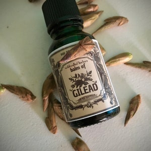 May include: A green glass bottle with a black cap, labeled "balm of GILEAD" in a vintage style. The bottle contains 1 fl oz of liquid. Scattered around the bottle are several brown seed pods. The label has a floral design and the words "wildcrafted tincture".