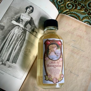 May include: A clear glass bottle with a black cap filled with a light yellow liquid. The bottle has a label with a red and gold design and the text "Milk of Frankincense" in black lettering. The label also includes a small illustration of a woman with long blonde hair and a floral design.