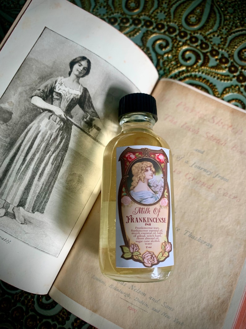 May include: A clear glass bottle with a black cap filled with a light yellow liquid. The bottle has a label with a red and gold design and the text "Milk of Frankincense" in black lettering. The label also includes a small illustration of a woman with long blonde hair and a floral design.