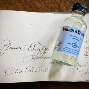 May include: A clear glass bottle with a black cap and a blue and white label that reads "Virgin's Milk" and "renders the skin clear and brilliant". The label also includes the year "1779" and the text "Beauty Jones". The bottle is sitting on a page of a book with handwritten text.