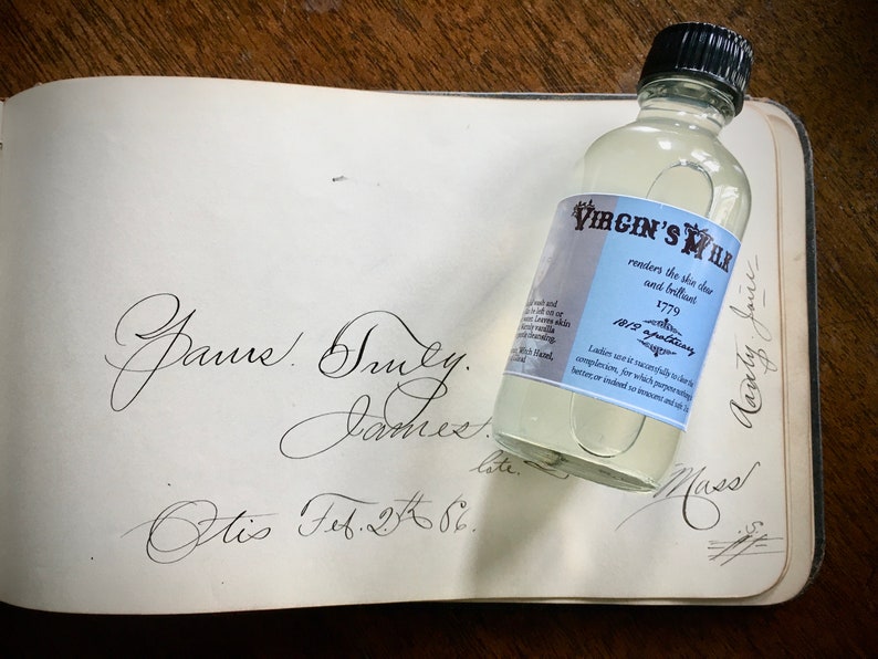 May include: A clear glass bottle with a black cap and a blue and white label that reads "Virgin's Milk" and "renders the skin clear and brilliant". The label also includes the year "1779" and the text "Beauty Jones". The bottle is sitting on a page of a book with handwritten text.