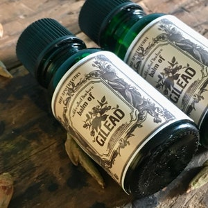May include: Two green glass bottles with black caps, labeled "Balm of Gilead" in a vintage style. The labels are cream-colored with floral and botanical illustrations. The bottles are on a rustic wooden surface.