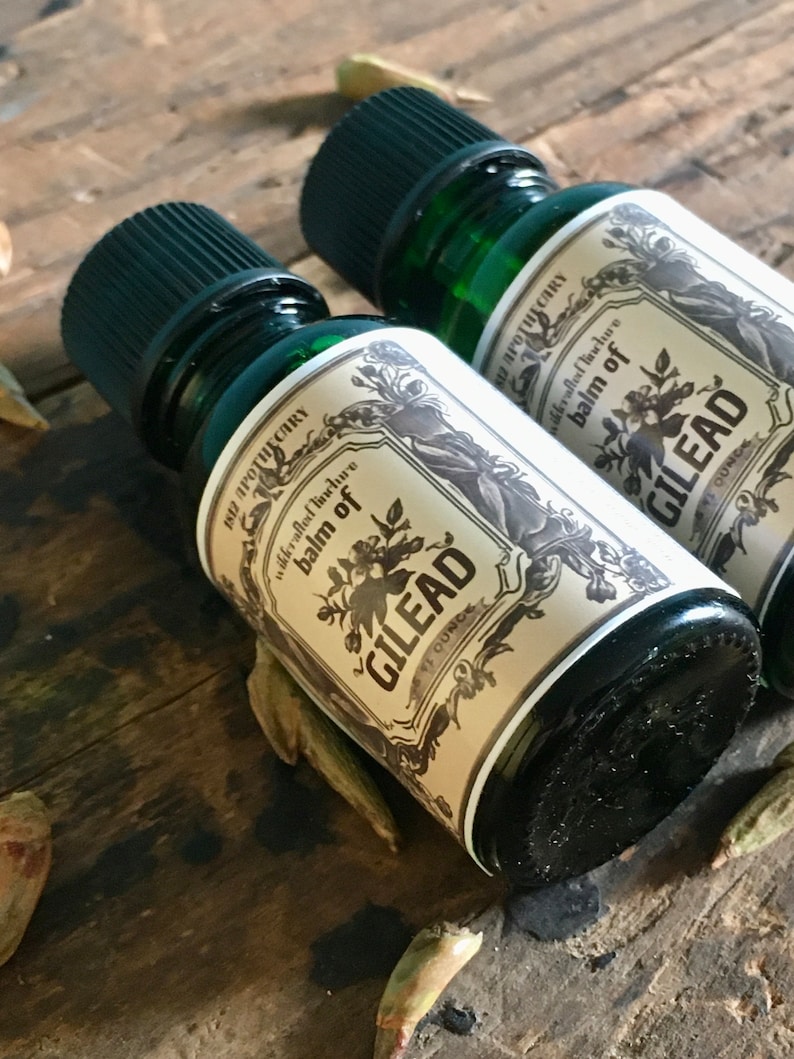 May include: Two green glass bottles with black caps, labeled "Balm of Gilead" in a vintage style. The labels are cream-colored with floral and botanical illustrations. The bottles are on a rustic wooden surface.