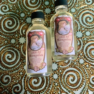 May include: Two small glass bottles with gold labels and black caps. The labels feature a vintage illustration of a woman and the text "Milk of Frankincense Oil".