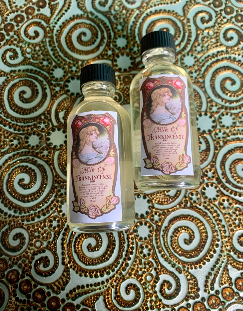 May include: Two small glass bottles with gold labels and black caps. The labels feature a vintage illustration of a woman and the text "Milk of Frankincense Oil".
