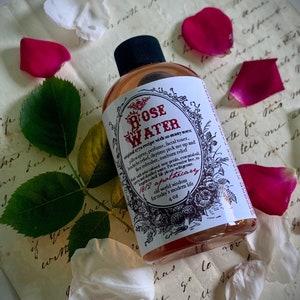 Op de afbeelding: Een doorzichtige glazen fles met een roze vloeistof erin. De fles heeft een wit etiket met zwarte tekst die "ROSE WATER original recipe with so many uses pillowspray, perfume, facial toner, headache relief, daytime pick me up and face refresher, sunburn relief Warwichlarinned rose petals, essential oil and distilled water. Preserative free, to lengthen shelf life please refrigerate. 1812 apothecary old world wisdom for today's modern life 4 OZ" luidt. De fles is omringd door rozenblaadjes.