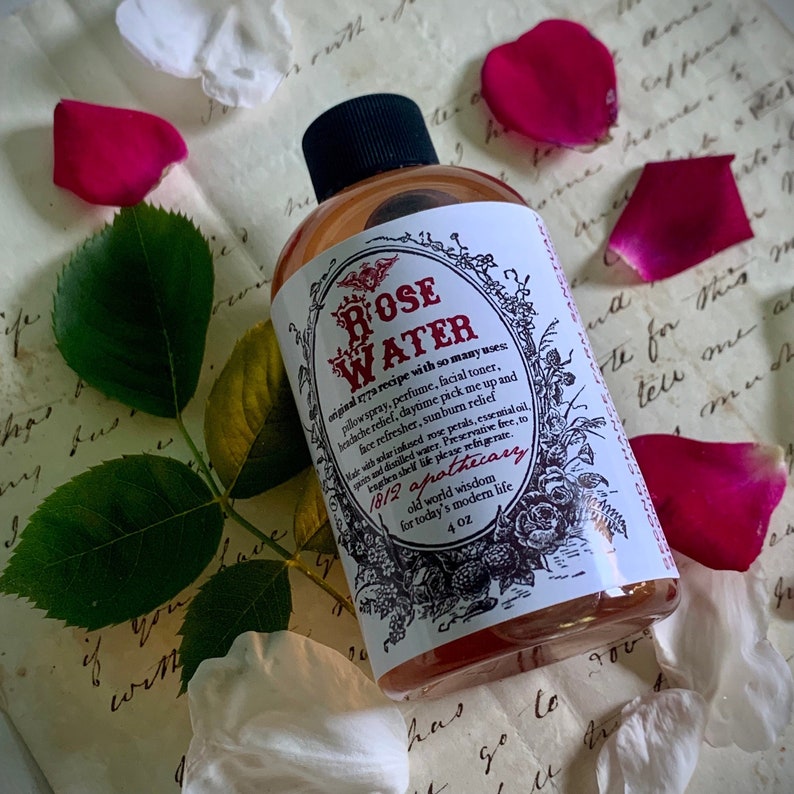 Op de afbeelding: Een doorzichtige glazen fles met een roze vloeistof erin. De fles heeft een wit etiket met zwarte tekst die "ROSE WATER original recipe with so many uses pillowspray, perfume, facial toner, headache relief, daytime pick me up and face refresher, sunburn relief Warwichlarinned rose petals, essential oil and distilled water. Preserative free, to lengthen shelf life please refrigerate. 1812 apothecary old world wisdom for today's modern life 4 OZ" luidt. De fles is omringd door rozenblaadjes.