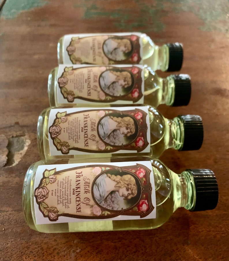 May include: Four small glass bottles with black caps and labels that say "Milk of Frankincense" in a vintage style font. The labels feature a woman in a white dress and a floral design.