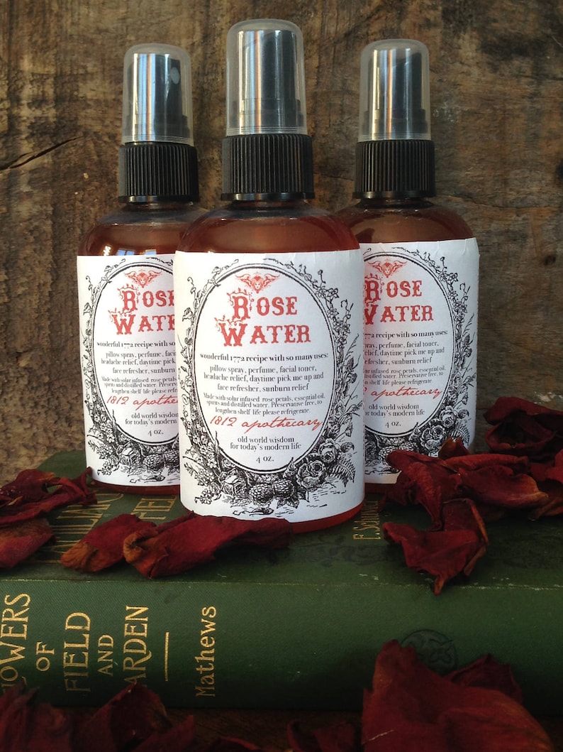 Op de afbeelding: Drie 4-ounce amber glazen flessen met zwarte spuitkoppen en witte etiketten. De etiketten hebben een vintage ontwerp en zeggen "Rose Water 1812 Apothecary old world wisdom for today's modern life". De flessen staan op een groen boek met rode rozenblaadjes eromheen.