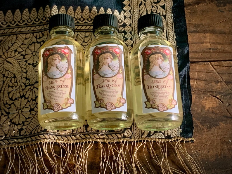 May include: Three clear glass bottles with gold labels and a vintage design. Each bottle contains a yellow liquid and is labeled "Milk of Frankincense". The bottles are sitting on a gold and black patterned fabric.
