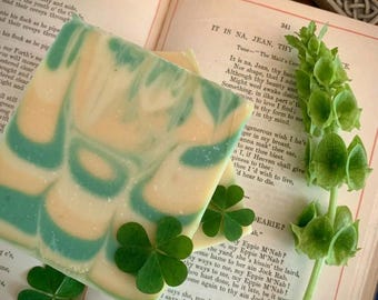 May include: A square bar of soap with a marbled pattern of cream and teal swirls. The soap is placed on an open book with printed text, accompanied by three green clover leaves and a tall green flower stalk with bell-shaped blossoms.