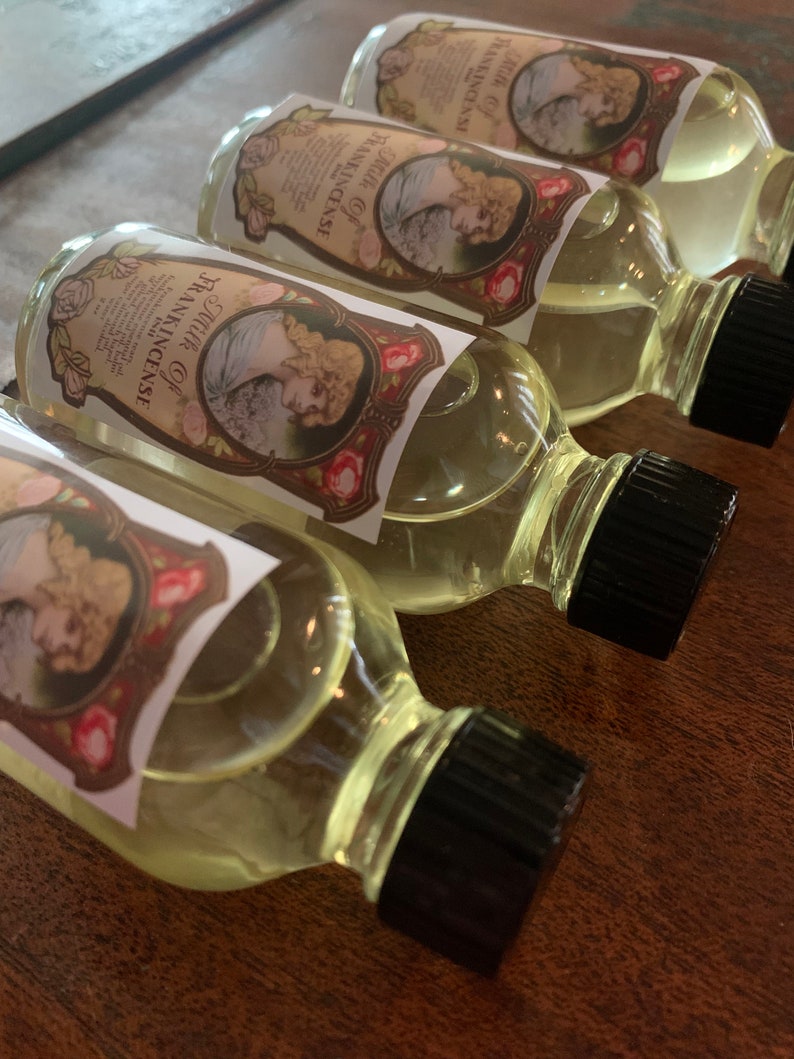 May include: Four clear glass bottles with black caps filled with a light yellow liquid. Each bottle has a label with a vintage design featuring a woman and roses. The label text reads "Pure Oil of Frankincense".