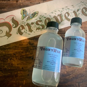 May include: Two clear glass bottles with white labels that read "The Virgin's Milk" and contain a clear liquid. The labels state that the liquid renders the skin clear and brilliant and is a 1770s antiquarian liqueur. The bottles are sitting on a wooden surface.