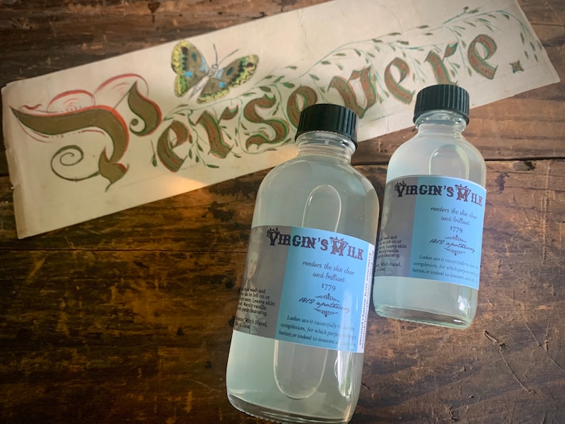 May include: Two clear glass bottles with white labels that read "The Virgin's Milk" and contain a clear liquid. The labels state that the liquid renders the skin clear and brilliant and is a 1770s antiquarian liqueur. The bottles are sitting on a wooden surface.
