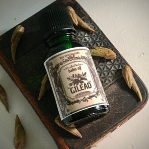 May include: A small, green glass bottle with a black cap and a cream-colored label that reads "balm of Gilead." The bottle sits on a dark, textured surface, surrounded by dried seed pods. The label also includes the text "1812 Apothecary."