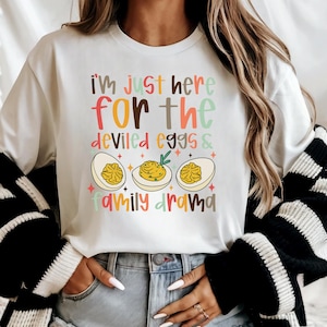 May include: White long-sleeve shirt with the text "I'm just here for the deviled eggs & family drama." The graphic includes three deviled eggs with colorful text and star accents. The shirt is a casual, comfortable style.