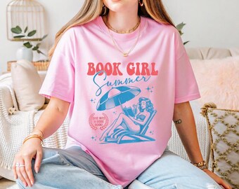 May include: Pink t-shirt with the words "BOOK GIRL Summer" in red and blue. The graphic features a woman reading under a beach umbrella. The text "Dive into a good book" is also included.