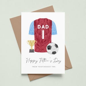 May include: A Father's Day card featuring a watercolor illustration of a burgundy and blue soccer jersey with the word "DAD" and the number "1". A gold trophy and a soccer ball are also depicted. The card reads "Happy Father's Day FROM YOUR BIGGEST FAN".