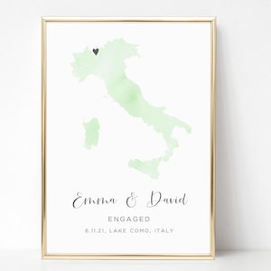 May include: A framed print featuring a light green watercolor map of Italy with a small black heart icon. Below the map, the text reads: Emma & David, ENGAGED, 6.11.21, LAKE COMO, ITALY. The print is enclosed in a thin gold-colored metal frame.