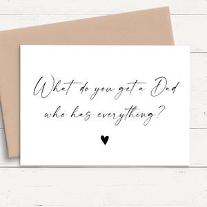May include: A white greeting card with black cursive text that reads "What do you get a Dad who has everything?" and a small black heart icon centred below the text, accompanied by a tan envelope.