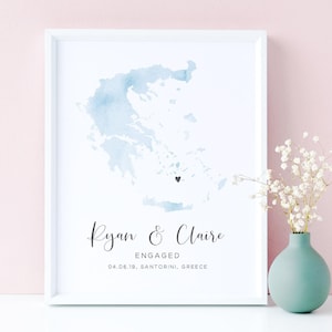 May include: A framed print featuring a light blue watercolor map of Greece with a small black heart marking a location. Below the map, the text reads Ryan & Claire, ENGAGED, and 04.06.19, SANTORINI, GREECE in black lettering.