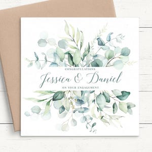 May include: A square engagement card featuring watercolor eucalyptus leaves in shades of green and sage. The text reads: CONGRATULATIONS Jessica & Daniel ON YOUR ENGAGEMENT. The card is paired with a plain brown envelope.
