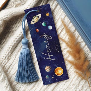 May include: A rectangular bookmark featuring a dark blue space-themed background with various planets, stars, and the name Henry written in white script. A light blue fabric tassel is attached to the top through a small circular hole.