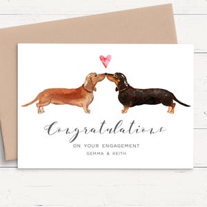 May include: A white greeting card featuring a watercolor illustration of a light brown and a dark brown dachshund touching noses under a small pink heart. The text reads Congratulations ON YOUR ENGAGEMENT GEMMA & KEITH in gray.