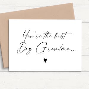 May include: A white greeting card with black cursive text that reads You're the best Dog Grandma... followed by a small black heart symbol. The card is positioned in front of a light brown envelope.