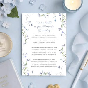 May include: A rectangular white greeting card featuring a blue floral border and the text To my Wife on your Heavenly Birthday, followed by a poem. The card is decorated with delicate blue flowers and green leaves printed on the paper.