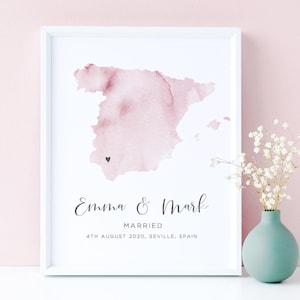 May include: A framed print featuring a pale pink watercolor map of Spain with a small black heart marking Seville. Below the map, the text reads Emma & Mark, MARRIED, 4TH AUGUST 2020, SEVILLE, SPAIN in black lettering on a white background.