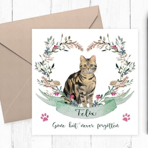 May include: A square greeting card featuring a watercolor illustration of a brown tabby cat centered within a floral wreath. A green ribbon banner below the cat displays the name Felix, with the text Gone but never forgotten and two pink paw prints.