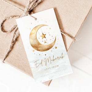May include: A gift tag with a watercolor design featuring a gold crescent moon, star, and the words "Eid Mubarak" in elegant script. The tag is tied to a brown paper package with twine.