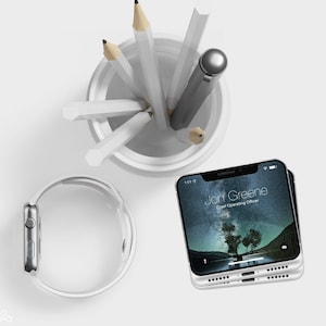 May include: A white Apple Watch with a white band, a white ceramic pencil holder with pencils and a pen, and a white iPhone with a black screen displaying the text "Jon Greene Chief Operating Officer" and a starry night sky background.