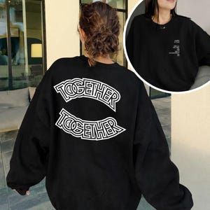 以下が含まれることがあります： 背中に白い「TOGETHER」の文字がアーチ状に描かれた黒いスウェットシャツ。その下に「TOGETHER」の文字が鏡面デザインで繰り返されています。小さな画像には、テキストが書かれたスウェットシャツの前面が写っています。