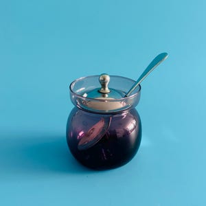May include: A small, round, purple glass jar with a clear lid and a silver spoon. The jar has a silver knob on the lid. The jar is on a blue background. The jar is likely for serving condiments or preserves.