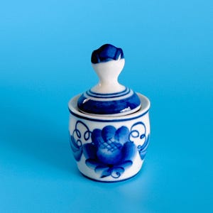 May include: A small white ceramic jar with a lid, featuring hand-painted blue floral patterns and swirling vine designs. The lid has a rounded knob handle, and the body of the jar is cylindrical with a slightly tapered base.