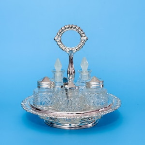 May include: A silver-plated condiment set with a handle, featuring a round tray, two glass jars with silver lids, and two glass bottles with stoppers. The set is on a blue background.