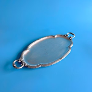 May include: An oval silver-toned serving tray with a smooth central surface and a decorative, scalloped rim. The tray features two ornate, circular handles on opposite ends, each detailed with intricate scrollwork and leaf-like patterns.