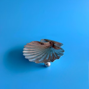 May include: A silver-toned metal dish shaped like a scallop shell with a ribbed surface texture. The shell dish features a small, spherical metal ball attached to the underside, serving as a singular support base for the object.