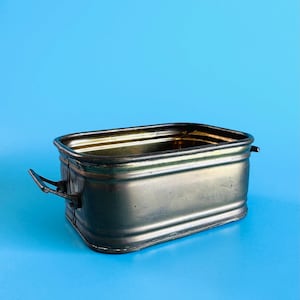 May include: A rectangular, silver-toned metal planter with two small handles on the sides. The planter has a slightly aged appearance and sits against a solid blue background. The container is empty.