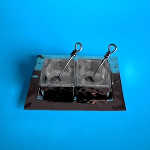 May include: A clear glass salt and pepper set with two square containers and small spoons. The set sits on a shiny, rectangular silver tray. The spoons have a looped design at the top. The background is a solid blue.