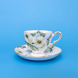 May include: A white teacup and saucer set with a floral pattern. The cup and saucer feature white flowers and green leaves, with gold edging. The teacup has a curved handle. The set is displayed against a blue backdrop.