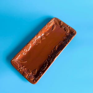 May include: A rectangular, brown ceramic serving tray with embossed leaf and acorn designs around the edges. The tray is set against a bright blue background, highlighting its decorative details and earthy tones. Suitable for serving appetizers or displaying decorative items.