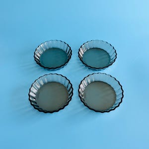 May include: A set of four round, translucent dark grey glass tart tins arranged in a two-by-two grid. Each tin features a fluted, scalloped edge design and a smooth, flat base, providing a consistent look for baking or serving individual portions.