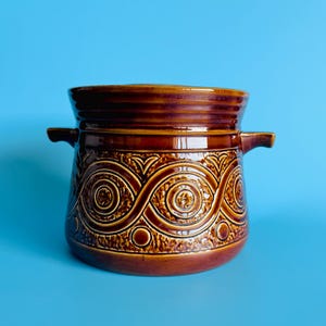 May include: A brown glazed ceramic pot with two side handles and a lid. The exterior features a repeating embossed pattern of interlocking circles and waves, with a smooth rim and a textured base section, showcasing a glossy finish throughout.