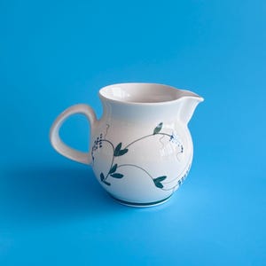 May include: A white ceramic cream jug featuring a rounded body, a curved handle, and a small pouring spout. The exterior is decorated with a delicate pattern of thin, dark green vines, small dark green leaves, and clusters of tiny blue flowers.