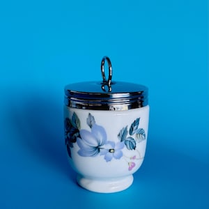 May include: A white porcelain egg cup with a silver lid and handle. The cup is decorated with a floral pattern in blue and green. The egg cup is set against a blue backdrop.