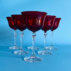 May include: Five ruby red stemmed glasses with a tulip-shaped bowl and clear stems and bases. The glasses are arranged in a row against a bright blue background. The glasses are likely for serving wine or cocktails.
