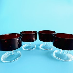 May include: Four clear glass dessert dishes with a dark red base. The dishes are arranged in a semi-circle on a light blue surface.